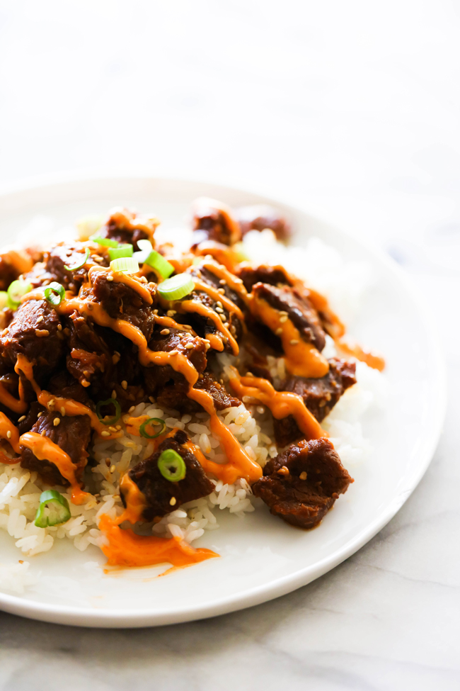 Korean Beef Bulgogi served on a white plate with Gochujang Mayo drizzled on top, garnished with green onions and sesame seeds.