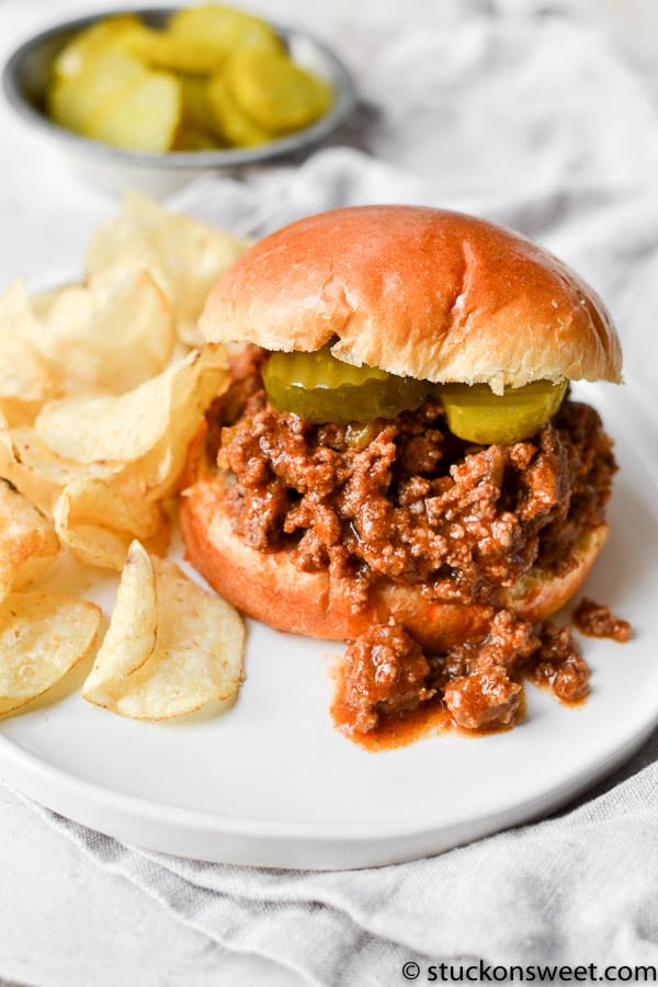 cooked ground beef meat on a bun with chips on a white plate.