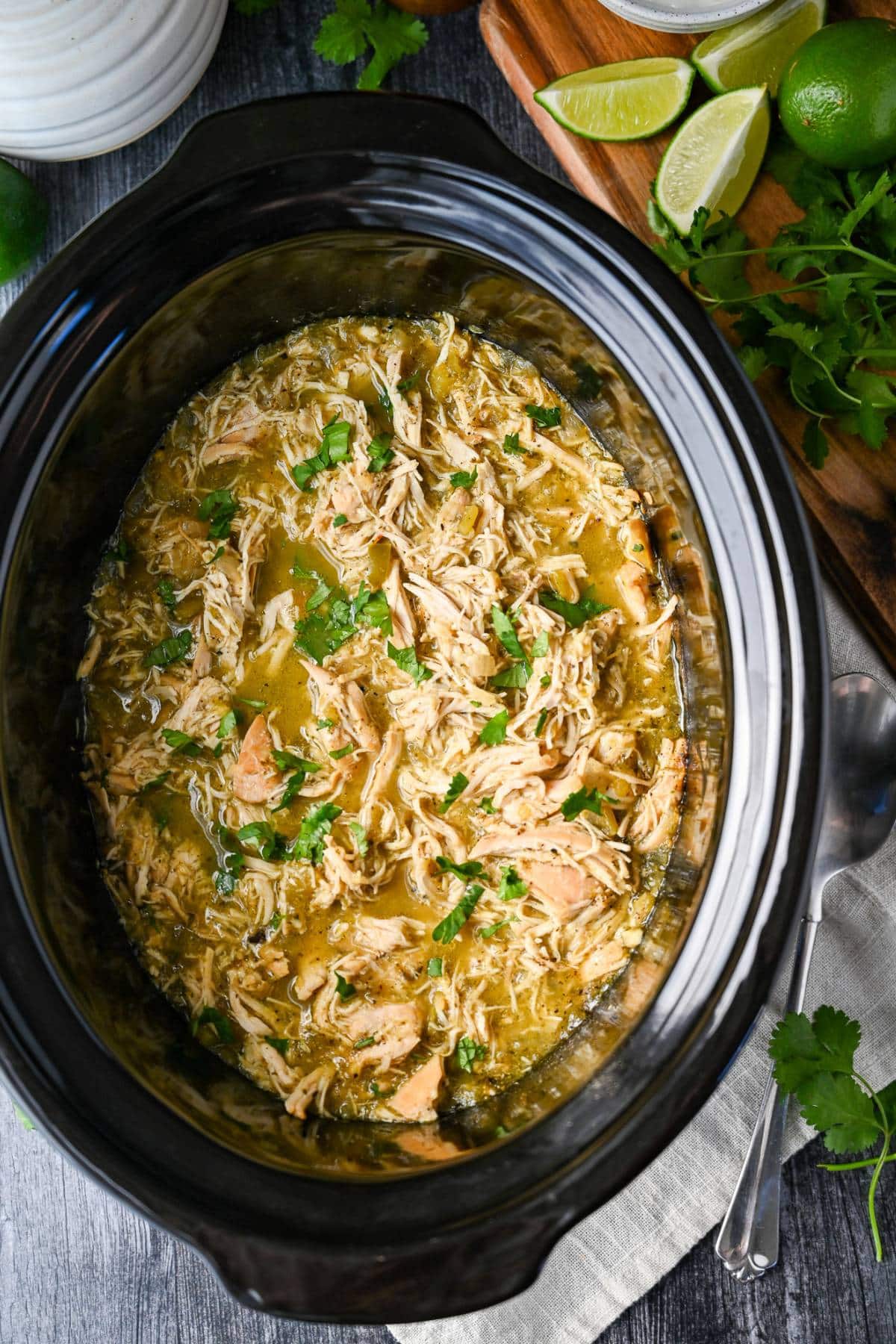 A view of a crockpot with shredded chicken simmering in a green chili sauce, garnished with fresh limes and cilantro, alongside a serving spoon