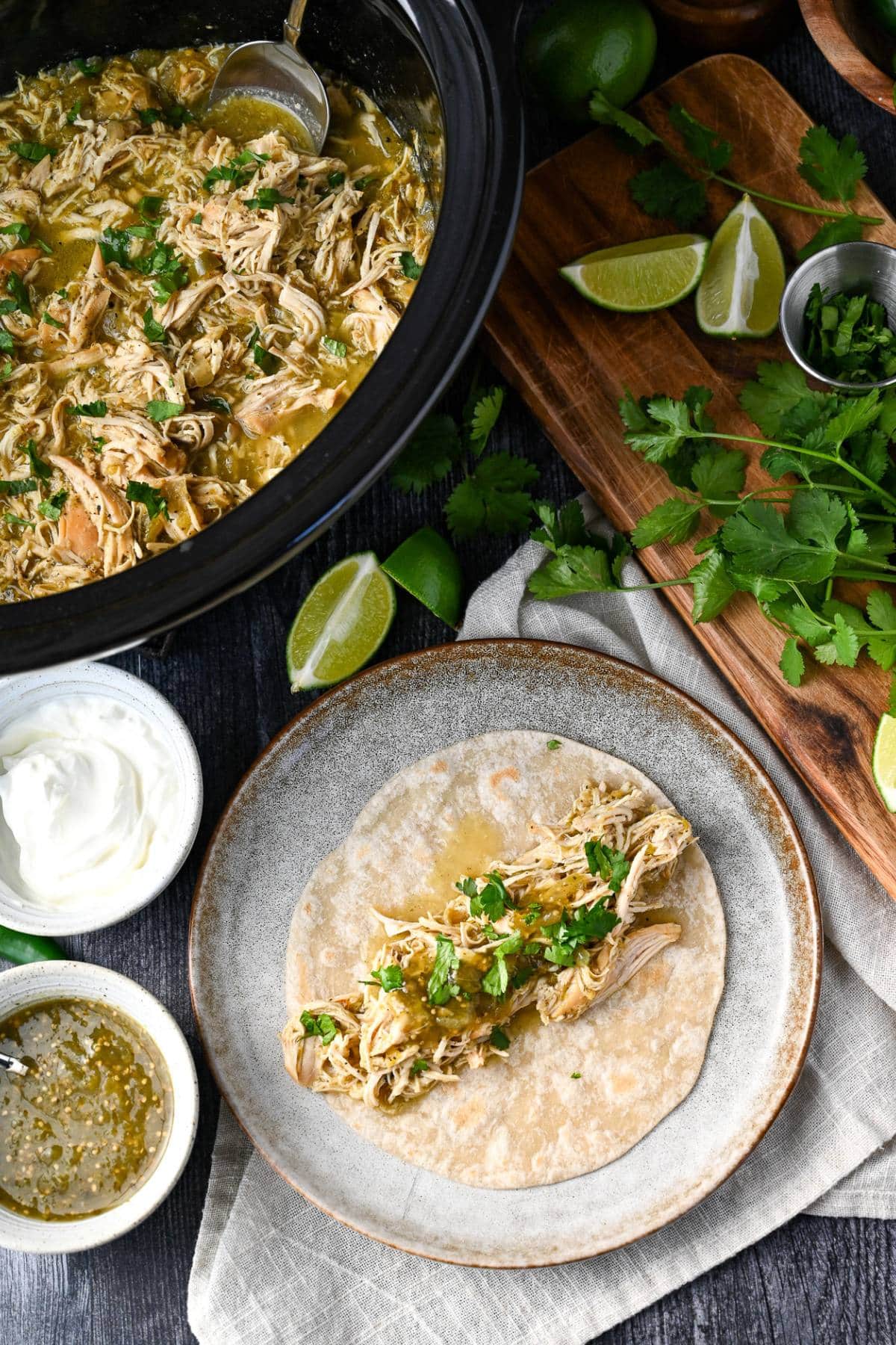 A tortilla topped with green chili shredded chicken, surrounded by garnishes and a slow cooker filled with the chicken behind it