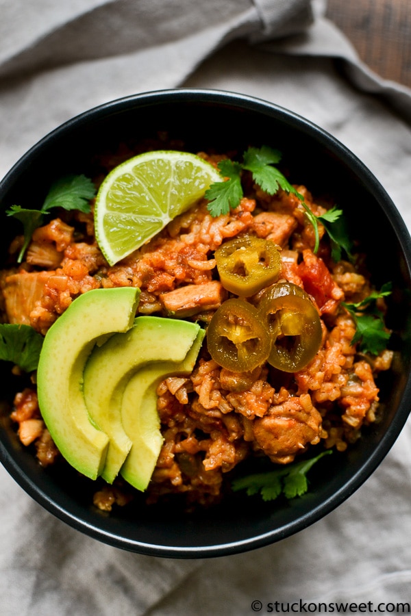 Close-up of Mexican rice in a black bowl topped with chicken, sliced avocado, lime wedge, and jalapeños.