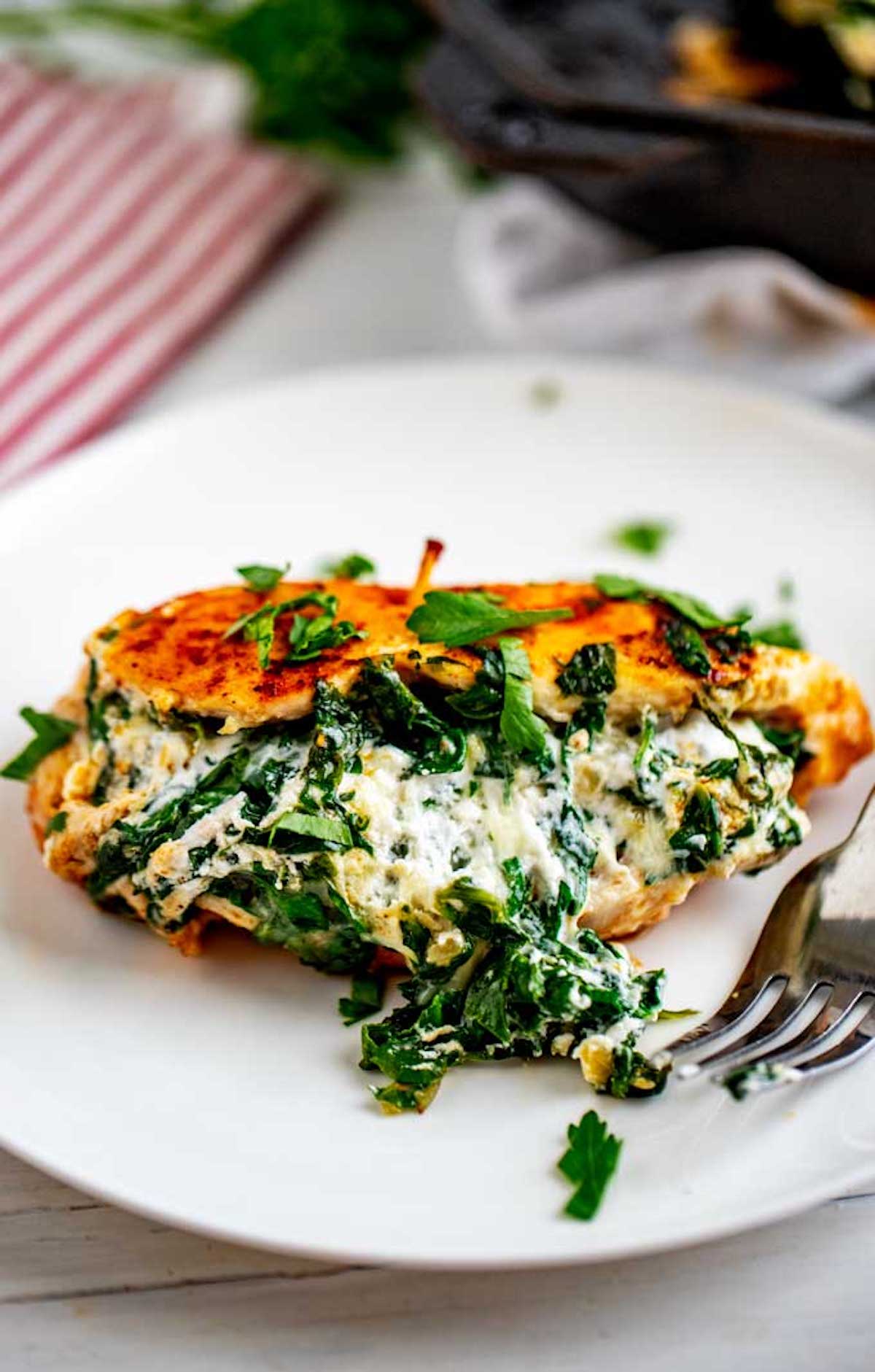 Image of a piece of Keto Stuffed Chicken on a white plate with cheese and spinach spilling out the front beside a fork.