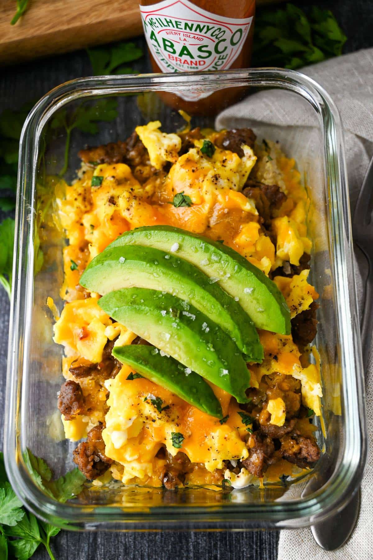 A breakfast bowl containing eggs, cheese, turkey sausage, and quinoa in a glass meal prep container, topped with avocado slices and accompanied by a bottle of hot sauce.