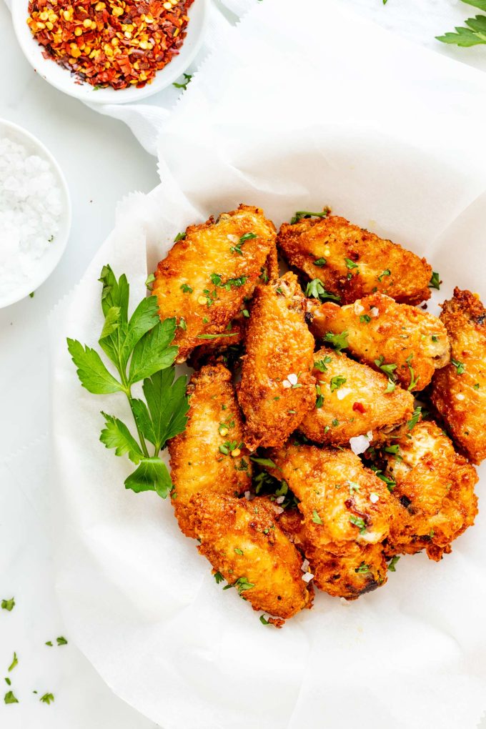 A top-down view of a parchment-lined plate filled with keto air fryer chicken wings alongside small bowls of Maldon sea salt and crushed red pepper flakes.