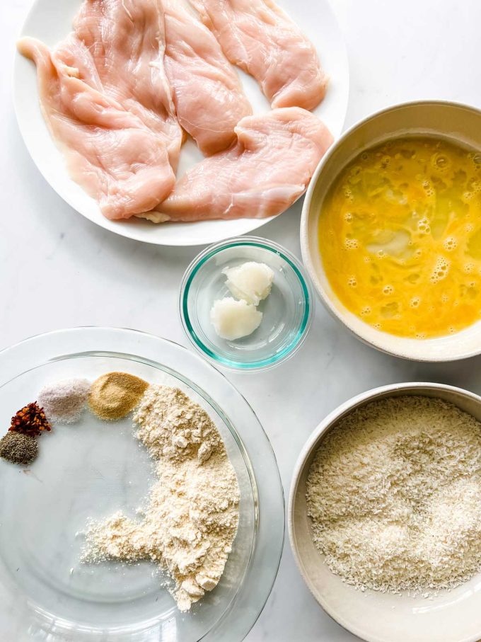 Overhead photo of chicken breast, beaten egg, coconut oil, coconut, coconut flour, and seasonings in prep bowls.