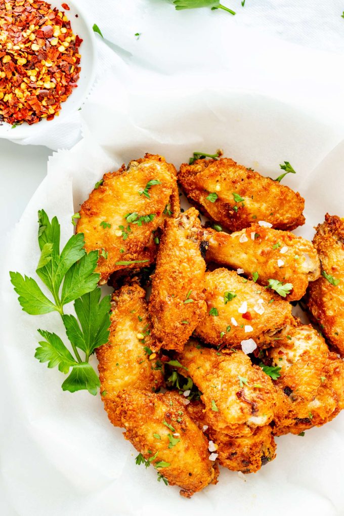 A top-down view of a parchment-lined plate filled with crispy keto air fryer chicken wings garnished with parsley, flaky salt, and crushed red pepper flakes.